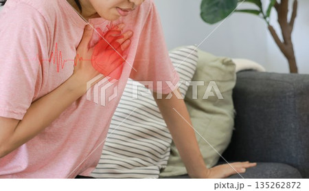 Asian woman sitting on sofa clutching chest with heart pain concept, healthcare, cardiology, emergency symptom, stress, medicine and heart attack awareness with ECG graphic overlay. 135262872