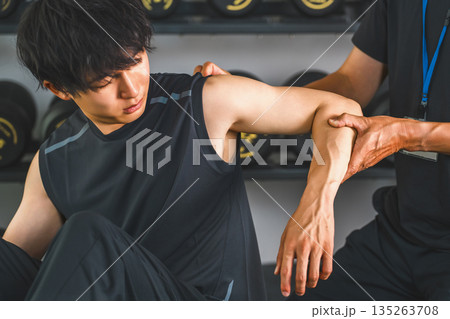 肩関節を痛がるトレーニングをする男性とスポーツ理学療法士の男性スタッフ 肩関節を痛がるトレーニングをする男性とスポーツ理学療法士の男性スタッフ 135263708