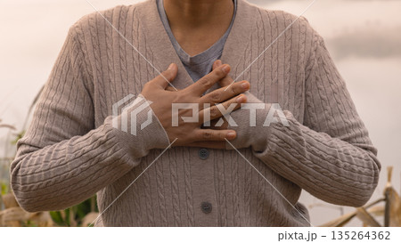 Close-up of a person with hands over heart in humble prayer, worshiping God with faith and gratitude. Warm knit sweater outdoors, spiritual devotion concept, copy space for church worship banners. Close-up of a person with hands over heart in humble prayer, worshiping God with faith and gratitude. Warm knit sweater outdoors, spiritual devotion concept, copy space for church worship banners. 135264362
