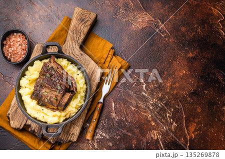 Close-up of tender beef short ribs, beautifully caramelized and resting on a bed of creamy mashed potatoes, creating an inviting culinary scene. brown background. top view Close-up of tender beef short ribs, beautifully caramelized and resting on a bed of creamy mashed potatoes, creating an inviting culinary scene. brown background. top view 135269878