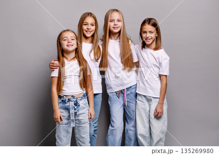 Group of happy children, preteen girls standing together in studio, smiling and looking at camera. Kids models in white t-shirts and jeans on gray background with copy space 135269980