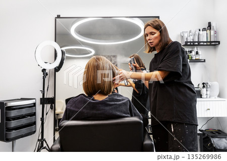 Hairstylist in modern salon styles chic bob haircut, applying hairspray for fixation and perfect hold. Professional beauty service in action Hairstylist in modern salon styles chic bob haircut, applying hairspray for fixation and perfect hold. Professional beauty service in action 135269986