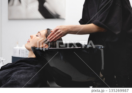 Hairdresser washing female clients hair with shampoo in black salon sink, professional beauty treatment while woman sits in black clothes inside modern studio Hairdresser washing female clients hair with shampoo in black salon sink, professional beauty treatment while woman sits in black clothes inside modern studio 135269987