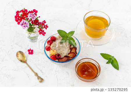 Oatmeal porridge with berries in a glass bowl and a cup of tea on the white table. 135270295