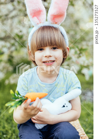 Easter Egg Hunt with Child Carrying Bright Plastic Eggs. Smiling child wearing bunny ears holding a wicker basket filled with colorful Easter eggs in a blooming spring garden 135273527