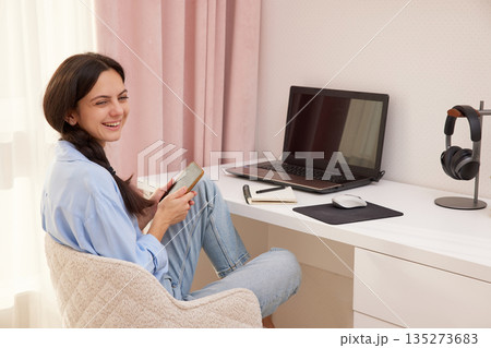 Happy woman holding a smartphone, sitting at a desk with a laptop 135273683