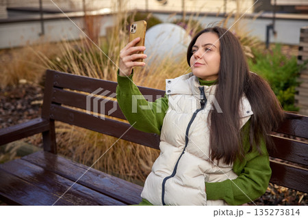 Young woman capturing a moment with her smartphone on a wooden bench outdoors Young woman capturing a moment with her smartphone on a wooden bench outdoors 135273814