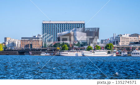 The Stockholm Waterfront Congress Centre stands prominently by the water, showcasing sleek modern design against a clear blue sky. Boats can be seen nearby on the tranquil waters. 135274571
