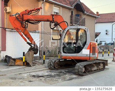Excavator working on residential street beside suburban houses at sunset. Road repair, urban infrastructure, and neighborhood development through construction activity and municipal service Excavator working on residential street beside suburban houses at sunset. Road repair, urban infrastructure, and neighborhood development through construction activity and municipal service 135275620