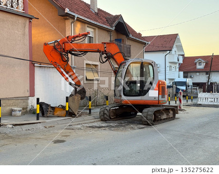 Excavator working on residential street beside suburban houses at sunset. Road repair, urban infrastructure, and neighborhood development through construction activity and municipal service Excavator working on residential street beside suburban houses at sunset. Road repair, urban infrastructure, and neighborhood development through construction activity and municipal service 135275622
