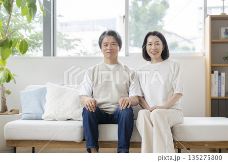 ソファに並んで座るカメラ目線で笑顔の50代の夫婦　イメージ 135275800