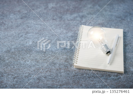 Glowing light bulb representing creative idea knowledge concept book, learn education learning innovation business training rests upon spiral notebook with pen over stone surface Glowing light bulb representing creative idea knowledge concept book, learn education learning innovation business training rests upon spiral notebook with pen over stone surface 135276461