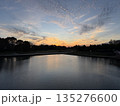 A spectacular sunset paints the sky with fiery oranges, reds, and yellows over a calm lake. The bright sun, low on the horizon, casts a brilliant reflection on the water. 135276600