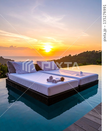 3D illustration of a luxury hotel bedroom with a comfortable bed and furniture facing a sunset over the sea through a large window 3D illustration of a luxury hotel bedroom with a comfortable bed and furniture facing a sunset over the sea through a large window 135276991