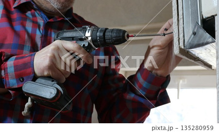 A worker in a plaid shirt, holding a black screwdriver, attaches a screw to a structure made of paneling and profiled metal indoors, creating an uneven partition in a room using a hand tool. A worker in a plaid shirt, holding a black screwdriver, attaches a screw to a structure made of paneling and profiled metal indoors, creating an uneven partition in a room using a hand tool. 135280959