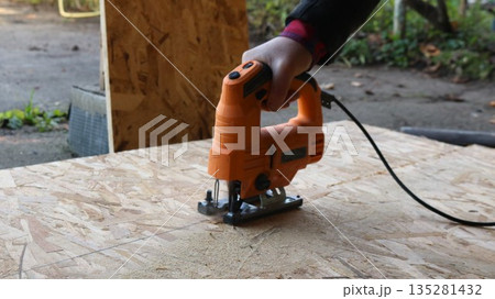 A worker's hand guides an orange electric jigsaw across an OSB board lying on the ground in a yard, sawing the sheet into two pieces. Cutting an OSB sheet with a jigsaw outdoors. 135281432