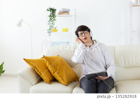Woman listening to music with headphones on sofa in modern living room. Audio relaxation, digital entertainment, and mindful leisure through home technology. 135282918