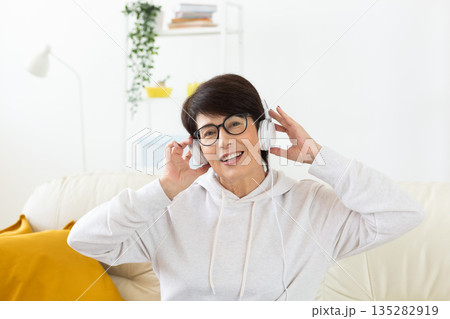 Woman listening to music with headphones on sofa in modern living room. Audio relaxation, digital entertainment, and mindful leisure through home technology. 135282919