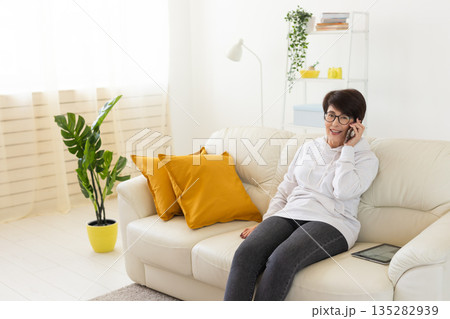 Woman talking on smartphone on white sofa in modern living room. Home communication, remote conversation, and digital lifestyle inside bright apartment interior. Woman talking on smartphone on white sofa in modern living room. Home communication, remote conversation, and digital lifestyle inside bright apartment interior. 135282939