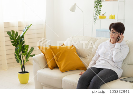 Woman talking on smartphone on white sofa in modern living room. Home communication, remote conversation, and digital lifestyle inside bright apartment interior. 135282941