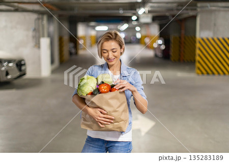 A woman smiles while holding a bag of fresh groceries in an underground parking garage 135283189