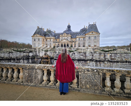 Maincy, France - December 11, 2025: Beautiful woman wearing a long red velvet cape, stands on an ornate stone bridge admiring the Chateau Vaux-le-Vicomte. Grand French Baroque Palace Maincy, France - December 11, 2025: Beautiful woman wearing a long red velvet cape, stands on an ornate stone bridge admiring the Chateau Vaux-le-Vicomte. Grand French Baroque Palace 135283523