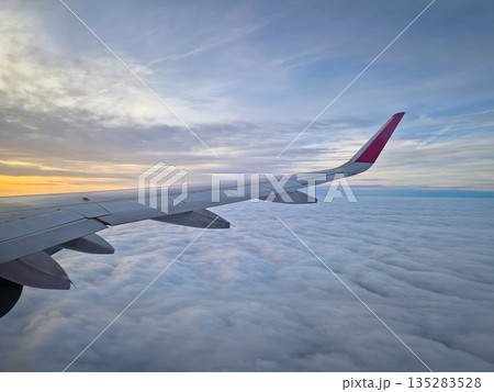 Peaceful aerial view from airplane window to the large wing soaring over a sea of clouds with the sun rising above the horizon, casting a warm glow and illuminating the cloudscape during flight 135283528