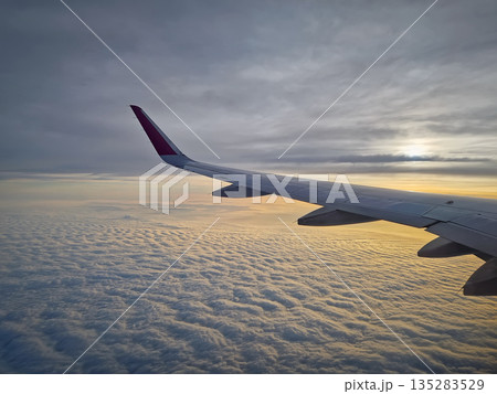 Peaceful aerial view from airplane window to the large wing soaring over a sea of clouds with the sun rising above the horizon, casting a warm glow and illuminating the cloudscape during flight 135283529