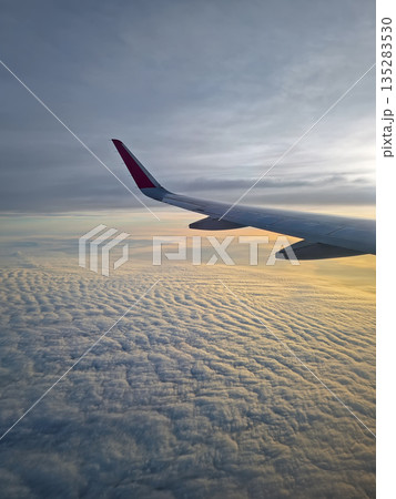 Peaceful aerial view from airplane window to the large wing soaring over a sea of clouds with the sun rising above the horizon, casting a warm glow and illuminating the cloudscape during flight 135283530