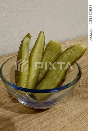 Crisp pickles sit in a glass bowl on a wooden table. The pickles are cut into long slices. The background is plain and neutral in color Crisp pickles sit in a glass bowl on a wooden table. The pickles are cut into long slices. The background is plain and neutral in color 135283688