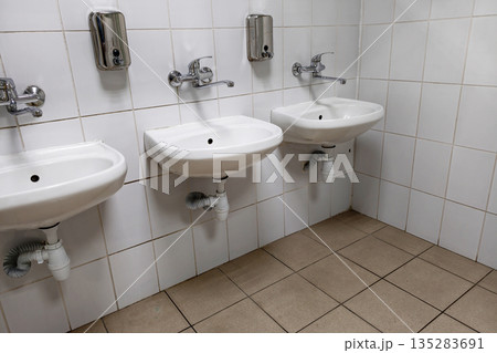 The restroom features three white sinks mounted on the wall. Each sink has a faucet and a soap dispenser installed above it The restroom features three white sinks mounted on the wall. Each sink has a faucet and a soap dispenser installed above it 135283691