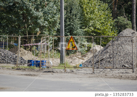 Workers are preparing a construction area marked by a fence and a warning sign. Dirt and materials are visible in the background among trees Workers are preparing a construction area marked by a fence and a warning sign. Dirt and materials are visible in the background among trees 135283733