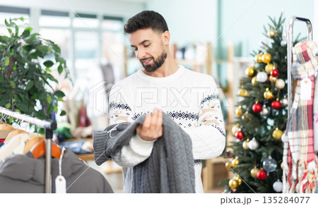 man chooses a sweater against the background of a Christmas tree in a store 135284077