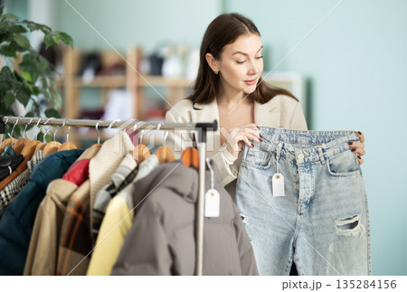 Happy young female client carefully choosing stylish jeans trousers in clothing store. Shopping and seasonal sales concept Happy young female client carefully choosing stylish jeans trousers in clothing store. Shopping and seasonal sales concept 135284156