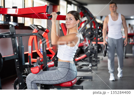 Girl doing strength training in gym, exercising upper body muscles on shoulder press machine 135284400