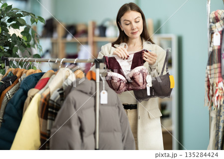 Focused young female client dressing in ordinary clothes holding bras in hands and selecting in underwear store Focused young female client dressing in ordinary clothes holding bras in hands and selecting in underwear store 135284424