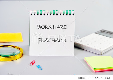A notepad on a desk says Work Hard Play Hard. Nearby are office supplies like a calculator, colorful paper clips, sticky notes, and a magnifying glass, suggesting a workspace A notepad on a desk says Work Hard Play Hard. Nearby are office supplies like a calculator, colorful paper clips, sticky notes, and a magnifying glass, suggesting a workspace 135284438