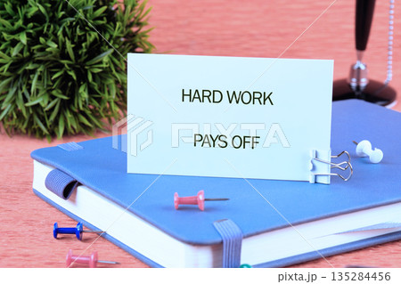 A card with the message HARD WORK PAYS OFF sits on a closed blue journal, surrounded by colorful push pins, an ornamental plant, and a writing pen, evoking a sense of motivation A card with the message HARD WORK PAYS OFF sits on a closed blue journal, surrounded by colorful push pins, an ornamental plant, and a writing pen, evoking a sense of motivation 135284456
