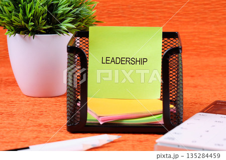 At the workspace, a green sticky note with the word Leadership sits inside a metal holder, next to a white plant pot and calculator. Various other colored notes are below At the workspace, a green sticky note with the word Leadership sits inside a metal holder, next to a white plant pot and calculator. Various other colored notes are below 135284459