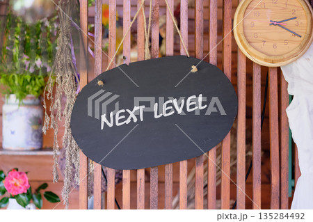 The black chalkboard with the white text Next Level is hanging on a wooden fence. A decorative clock and fresh flowers are visible in the background The black chalkboard with the white text Next Level is hanging on a wooden fence. A decorative clock and fresh flowers are visible in the background 135284492