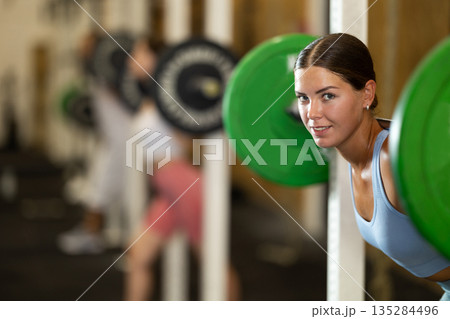 Motivated crossfit female athlete lifting heavy body bar or barbell during group training in sport gym Motivated crossfit female athlete lifting heavy body bar or barbell during group training in sport gym 135284496
