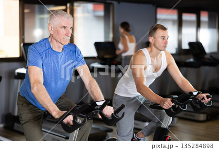 Two men exercising on stationary exercise bikes in gym Two men exercising on stationary exercise bikes in gym 135284898