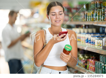 Girl scanning barcode or qr code with smartphone on sauce label in supermarket Girl scanning barcode or qr code with smartphone on sauce label in supermarket 135284973