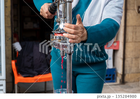 A woman extracts pomegranate juice using a manual juicer. She has dark hair and wears a blue outfit. The scene is set in a market environment. A woman extracts pomegranate juice using a manual juicer. She has dark hair and wears a blue outfit. The scene is set in a market environment. 135284980