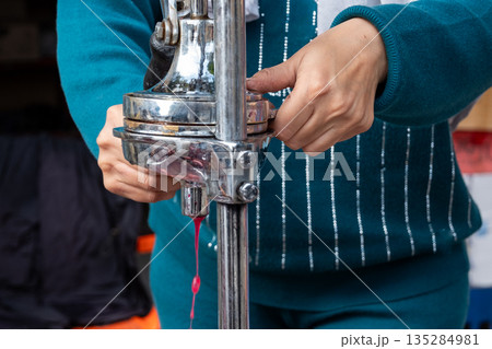 A woman extracts pomegranate juice using a manual juicer. She has dark hair and wears a blue outfit. The scene is set in a market environment. A woman extracts pomegranate juice using a manual juicer. She has dark hair and wears a blue outfit. The scene is set in a market environment. 135284981