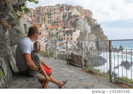 Father and young son sitting on a coastal path overlooking a seaside town. Family travel, bonding and lifestyle concept 135285336
