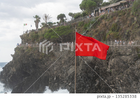 Red warning flag on a rocky seaside cliff above the sea. Concept of danger, storm, safety warning, strong waves and coastal conditions 135285338
