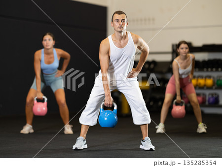 Sportive male in activewear swinging kettlebell during group exercise class in crossfit gym indoors Sportive male in activewear swinging kettlebell during group exercise class in crossfit gym indoors 135285354