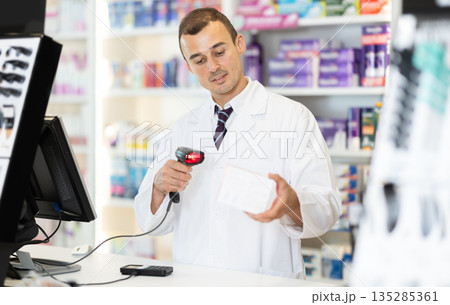 Man pharmacist in cash area works with computer and goods, scans drugs, checks availability Man pharmacist in cash area works with computer and goods, scans drugs, checks availability 135285361