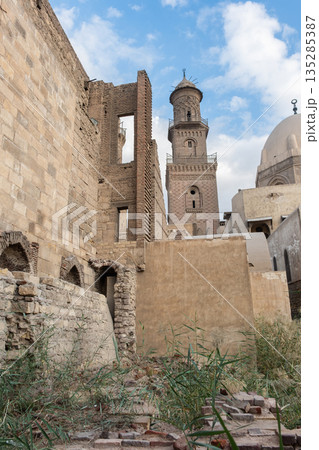 Historic Qalawun Complex in Cairo, featuring intricate architecture, tall minarets, and domes under a blue sky with clouds. A significant cultural landmark on Al-Muizz Street. Historic Qalawun Complex in Cairo, featuring intricate architecture, tall minarets, and domes under a blue sky with clouds. A significant cultural landmark on Al-Muizz Street. 135285387